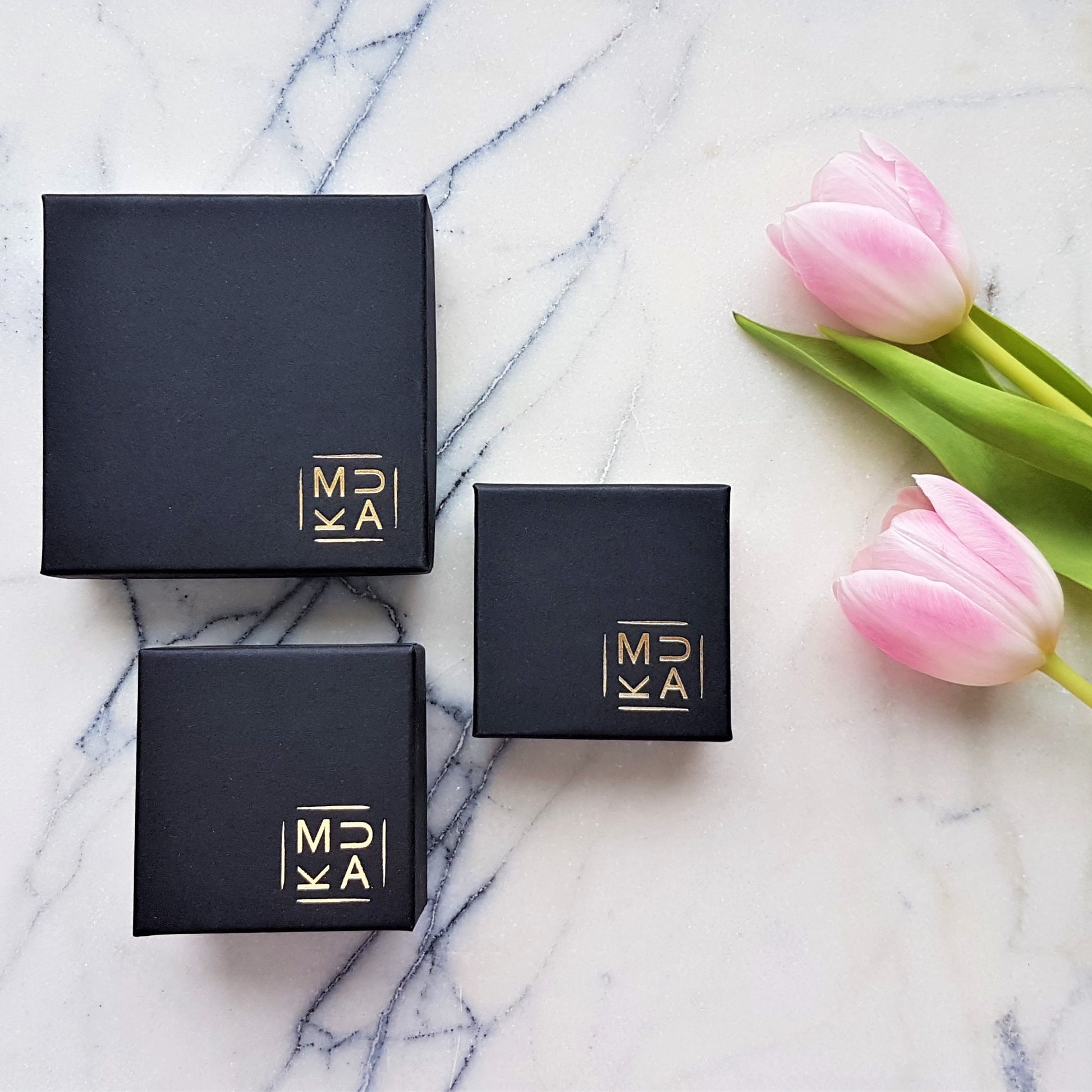 MUKA studio packaging, black jewellery boxes with gold logo placed on marble tile next to pink tulip flowers