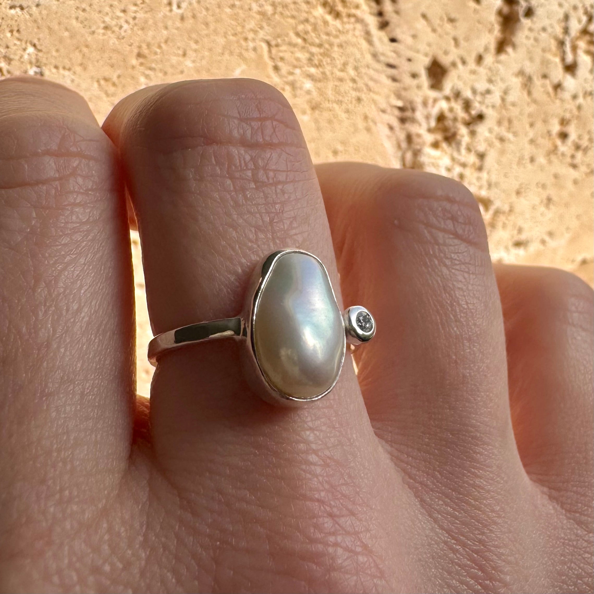 Silver ring with a large pearl stone and diamond on a finger against a beige background