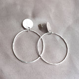 A pair of silver hoop earrings with flat circular disc charms, displayed on a textured gray fabric background.
