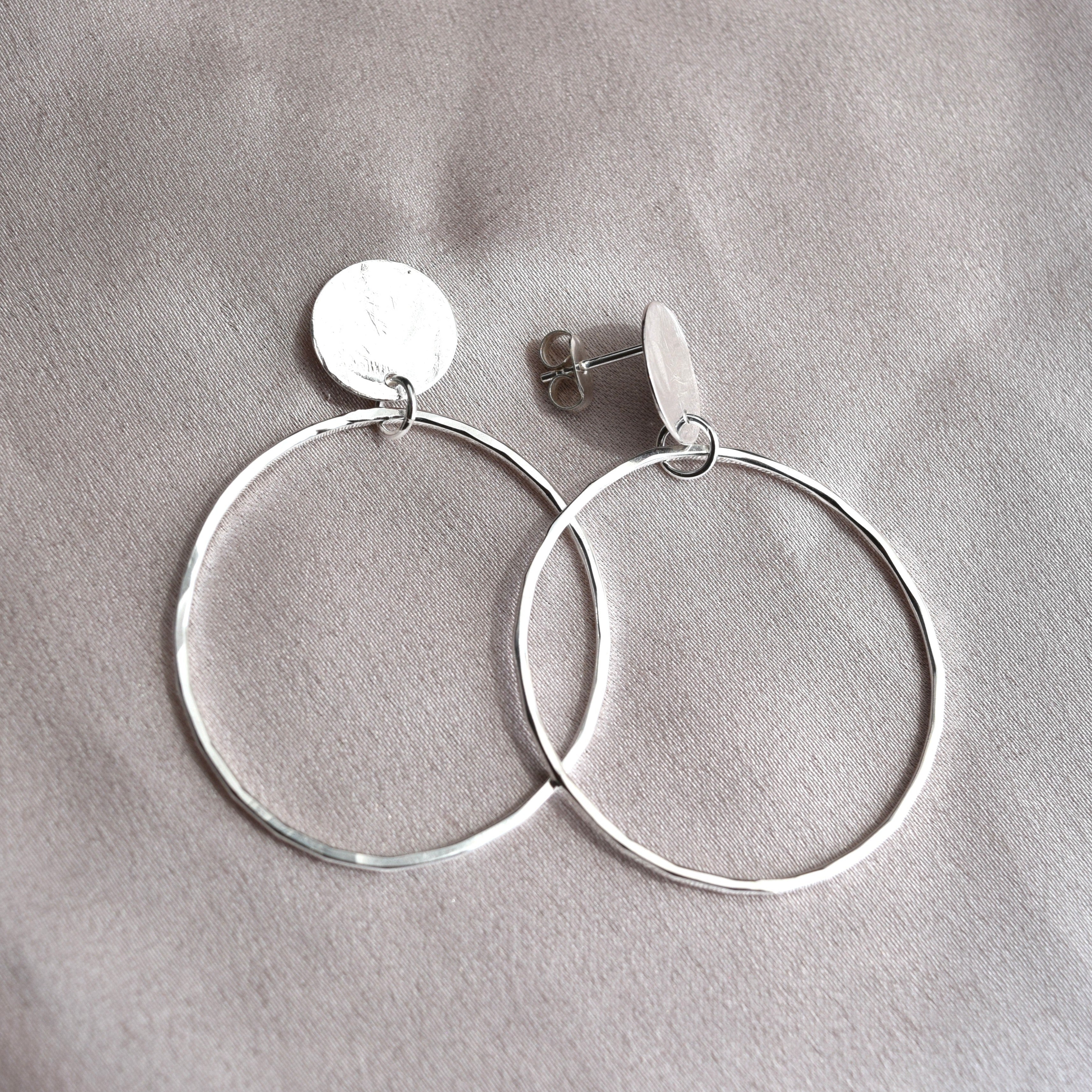 A pair of silver hoop earrings with flat circular disc charms, displayed on a textured gray fabric background.