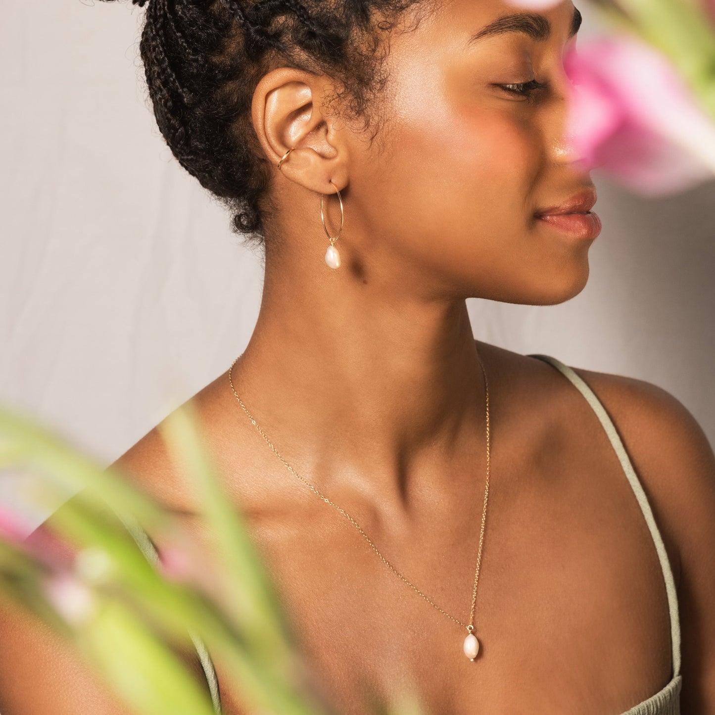 model wears gold pearl hoops, gold conch cuff and gold pearl necklace. Image is shot with pink flowers in the foreground slightly out of focus and model in the background in focus
