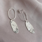 A pair of silver dangle earrings with hammered oval leaf-shaped charms, displayed on a soft, draped light gray fabric background.
