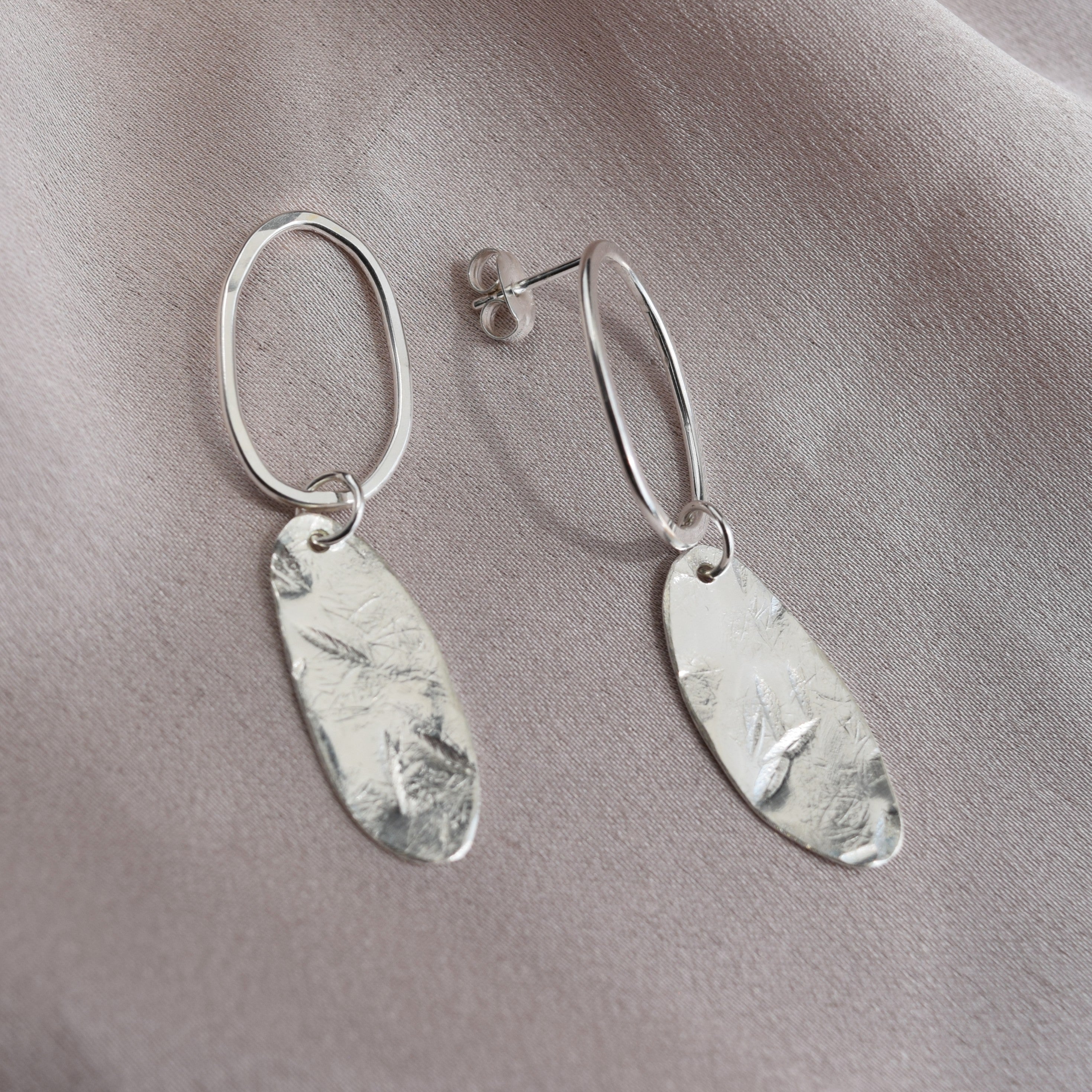 A pair of silver dangle earrings with hammered oval leaf-shaped charms, displayed on a soft, draped light gray fabric background.
