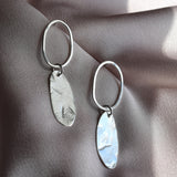 A pair of silver drop earrings with oval hoops and textured, hammered oval pendants, displayed on draped satin fabric.