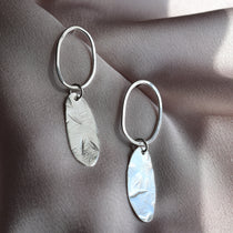 A pair of silver drop earrings with oval hoops and textured, hammered oval pendants, displayed on draped satin fabric.