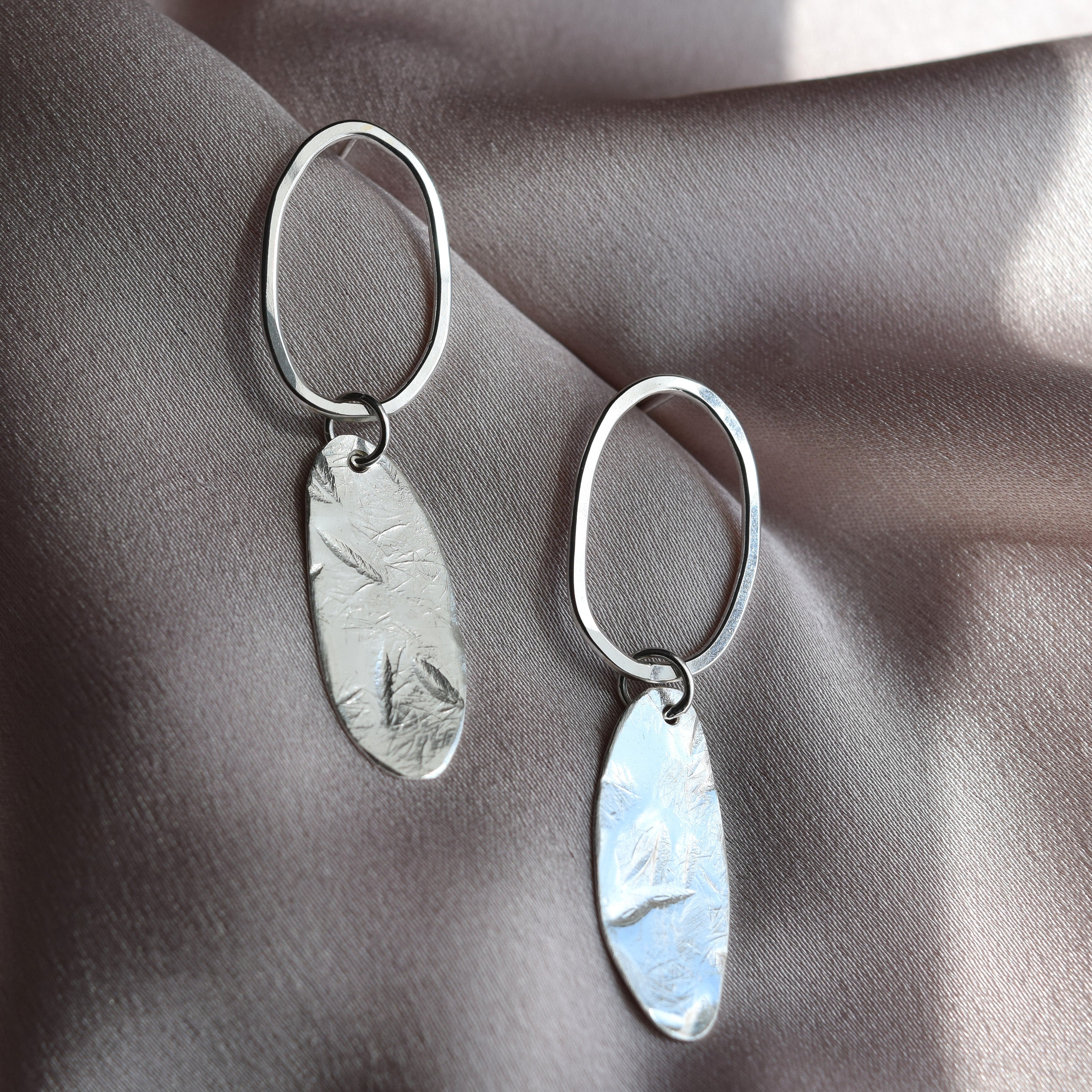 A pair of silver drop earrings with oval hoops and textured, hammered oval pendants, displayed on draped satin fabric.