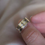 A close-up of a hand wearing a multi-band ring with alternating gold and silver metallic bands, resting against a soft beige fabric background.