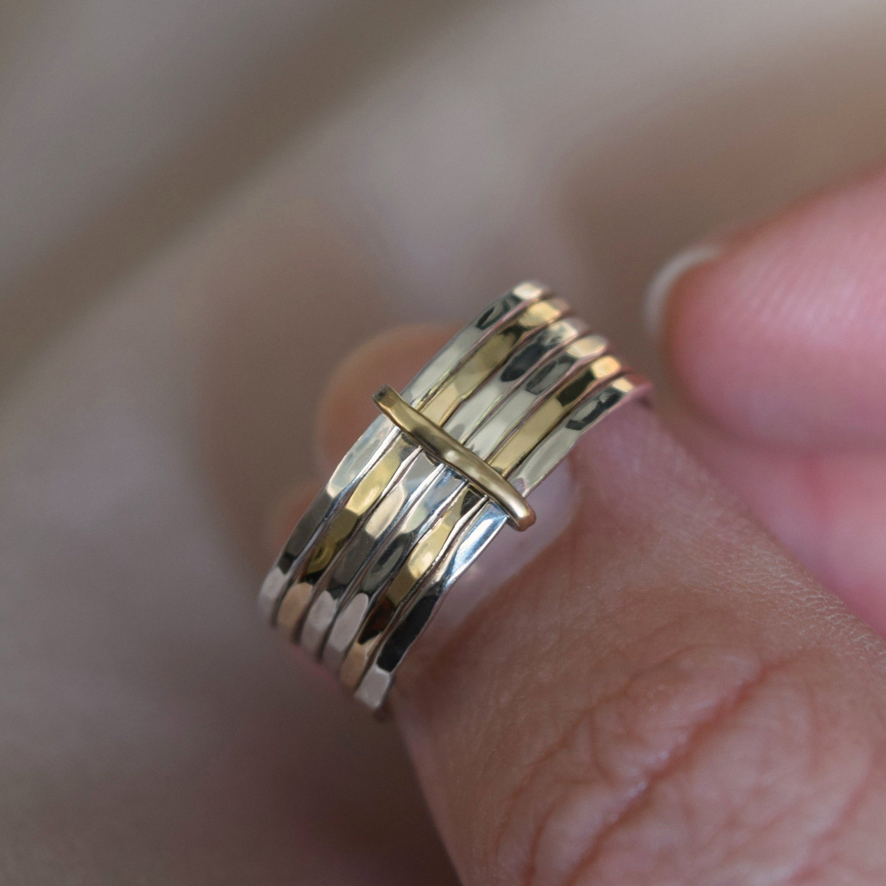 A close-up of a hand wearing a multi-band ring with alternating gold and silver metallic bands, resting against a soft beige fabric background.