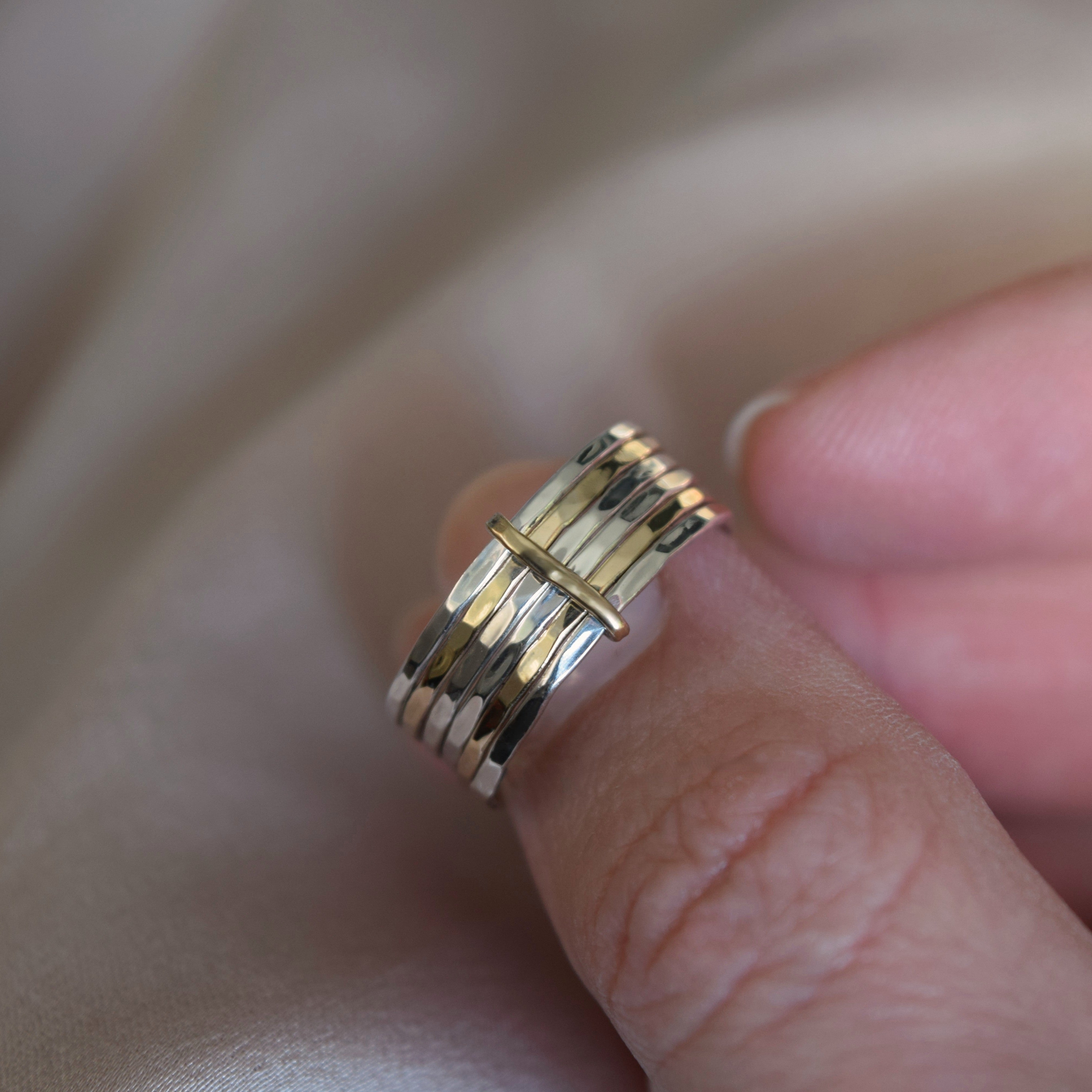 A close-up of a hand wearing a multi-band ring with alternating gold and silver metallic bands, resting against a soft beige fabric background.