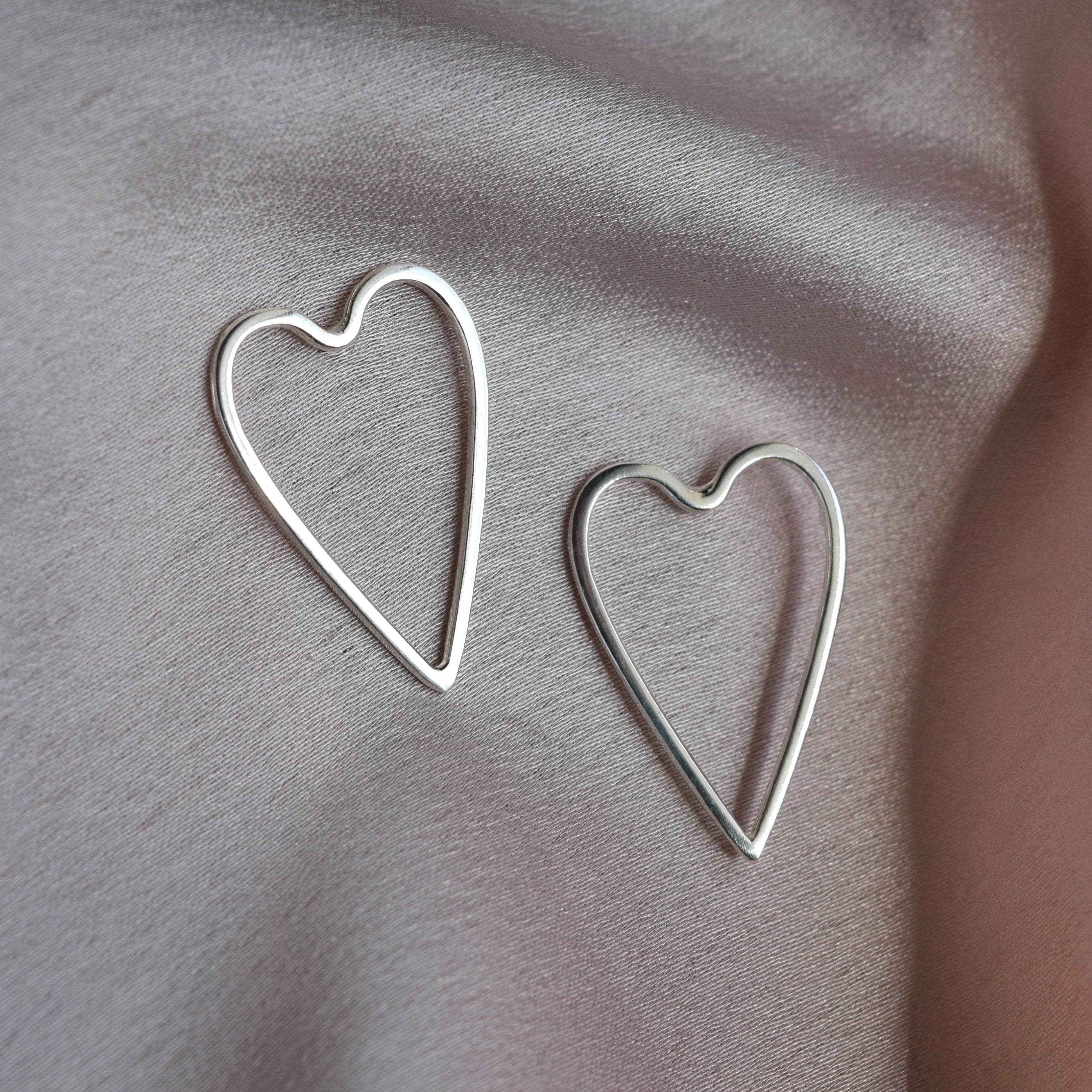 Two silver heart-shaped wire earrings displayed on a draped, light gray satin fabric background.