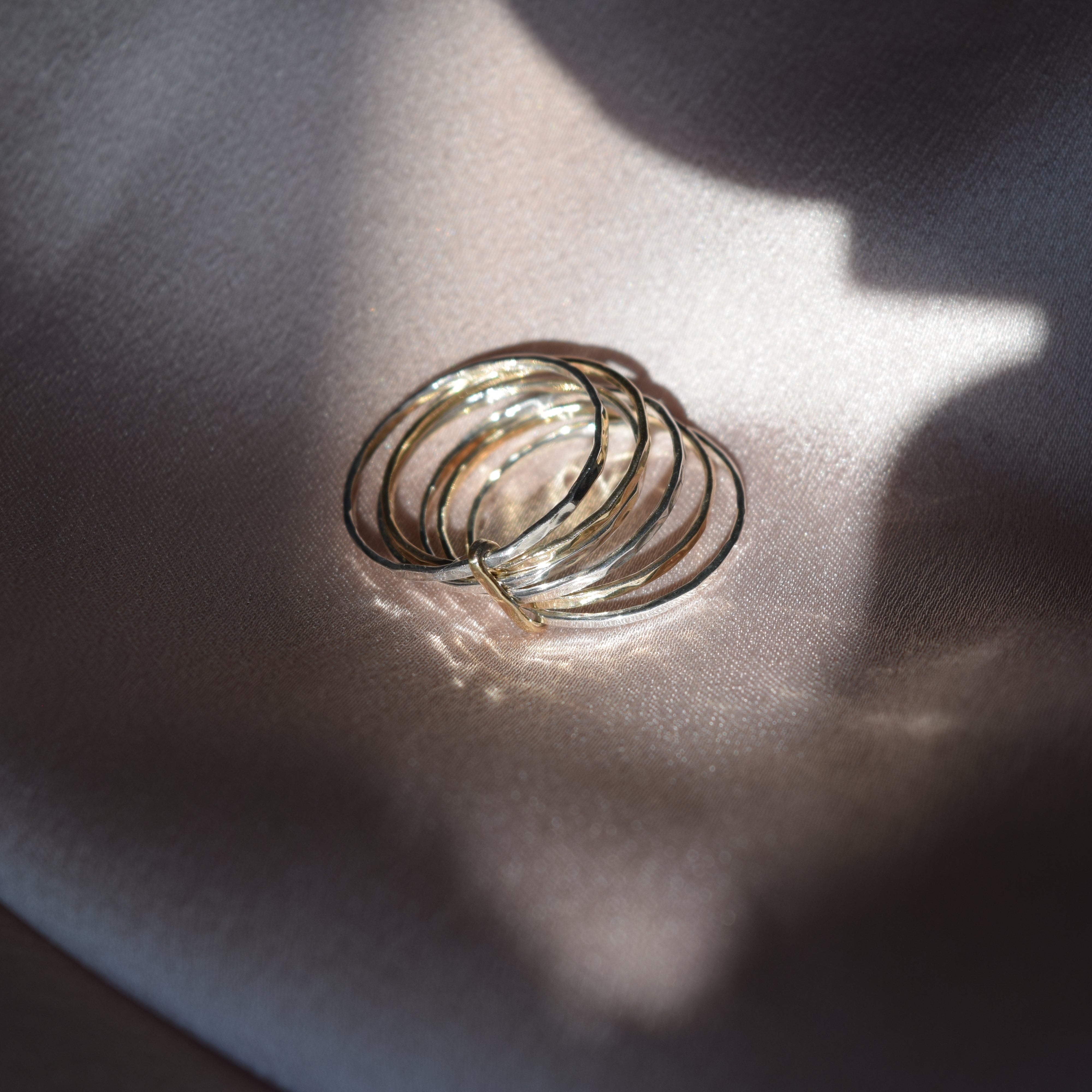 A stack of thin gold rings resting on a draped satin fabric under dramatic, directional sunlight and shadow.